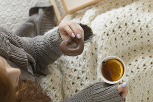 Person holding a cup of tea and a sugary snack keeping warm under a blanket